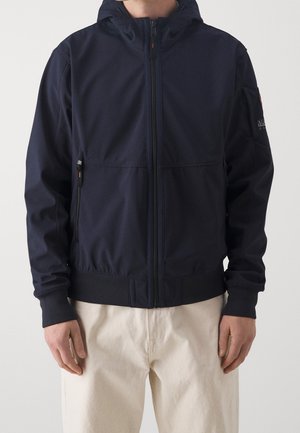 Mies pukeutunut laivastonsiniseen vetoketjulliseen huppariin, jossa sivutaskut, ja vaaleanbeigeihin housuihin yksivärisen taustan edessä.