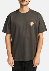 Dunkelgraues Baumwoll-T-Shirt mit einem kleinen orangen Stern-Grafik und türkisfarbenen Akzenten auf der linken Brust. Kurze Ärmel, Rundhalsausschnitt.