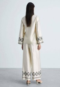 Cream-colored two-piece outfit featuring a long-sleeved top and wide-leg pants, adorned with black geometric embroidery on cuffs and hems.
