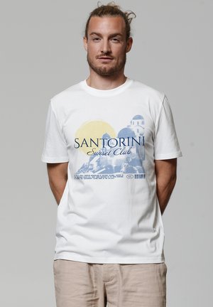 Man with light brown hair tied back wearing white Santorini Sunset Club t-shirt and beige pants, standing against plain gray background.
