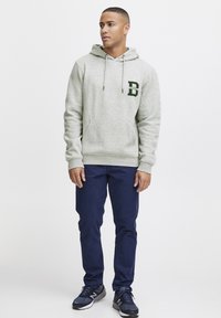 Light gray hoodie with front kangaroo pocket, drawstring hood, and green embroidered letter "B." Paired with dark blue pants and sports shoes.