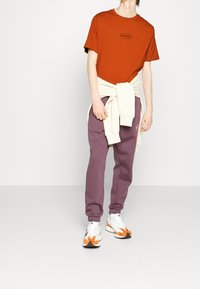 Rustkleurig T-shirt met een klein zwart logo, gecombineerd met een crèmekleurige trui die om de taille is gestrikt, gecombineerd met paarse trainingsbroek en kleurrijke sneakers.