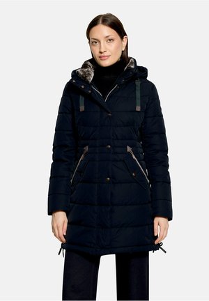 Femme portant un manteau d'hiver matelassé noir avec une capuche bordée de fourrure et des poches zippées, debout devant un fond clair uni.