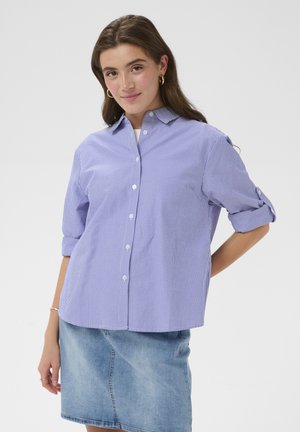 Frau trägt ein blau-weiß gestreiftes Hemd mit aufgerollten Ärmeln und einen hellblauen Jeansrock, posiert vor einem einfarbigen Hintergrund.