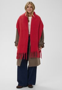 Red fringed wrap with a soft texture layered over a brown coat. The outfit includes a white top and wide-leg dark denim pants.