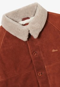 Chaqueta de ante marrón con un cuello de borreguillo beige, dos botones marrones y un pequeño logo bordado en dorado en el pecho.