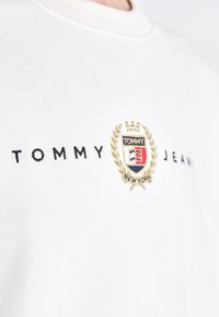 Balts kokvilnas džemperis ar apaļu izgriezumu, ar izšūtu logotipu zilā, zelta un sarkanā krāsā, kā arī lauru vainaga dizainu.