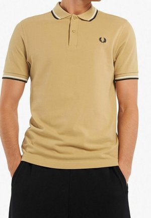 Mężczyzna w beżowej koszulce polo z krótkim rękawem, z czarnym i białym wykończeniem na kołnierzyku i rękawach, w czarnych spodniach, z rękami w kieszeniach.