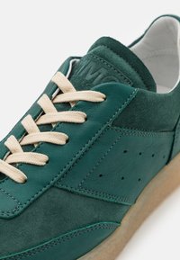 Tmavě zelené kožené tenisky s krémovými tkaničkami, semišovými akcenty, perforovanými stranami a texturovanou hnědou gumovou podrážkou. Logo je embosováno na jazyku.
