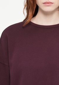Burgundské mikina s okrúhlym výstrihom, jemnou textúrou a uvoľneným strihom. Švy dodávajú celkovému dizajnu jemný záujem.