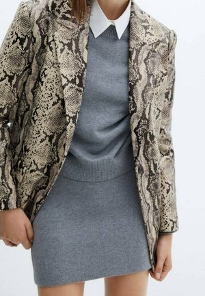 Vrouw die een grijze gebreide jurk draagt met een witte kraag onder een beige en zwarte blazer met slangenhuidpatroon.