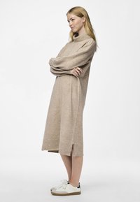 Robe en tricot beige avec un col montant, des manches longues, des fentes latérales et des poignets côtelés, associée à des baskets blanches avec une touche foncée.