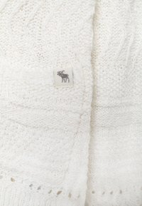 Biały sweter dzianinowy z wzorem w warkocze, ściągaczami przy mankietach oraz małą metką z szarym wzorem plecaka.