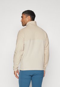 Beige fleecejakke med høj krave og lange ærmer, der har et kontrasterende struktureret panel over den øverste del af ryggen.