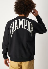Persona con cabello rizado que lleva una sudadera negra con "CHAMPION" en letras grandes blancas en la espalda, combinada con pantalones color beige.