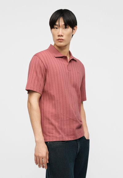 Νέος άνδρας με ίσια μαύρα μαλλιά, φορώντας ροζ πόλο μπλουζάκι με κοντά μανίκια και σκούρο τζιν, στέκεται μπροστά σε απλό ανοιχτό φόντο.