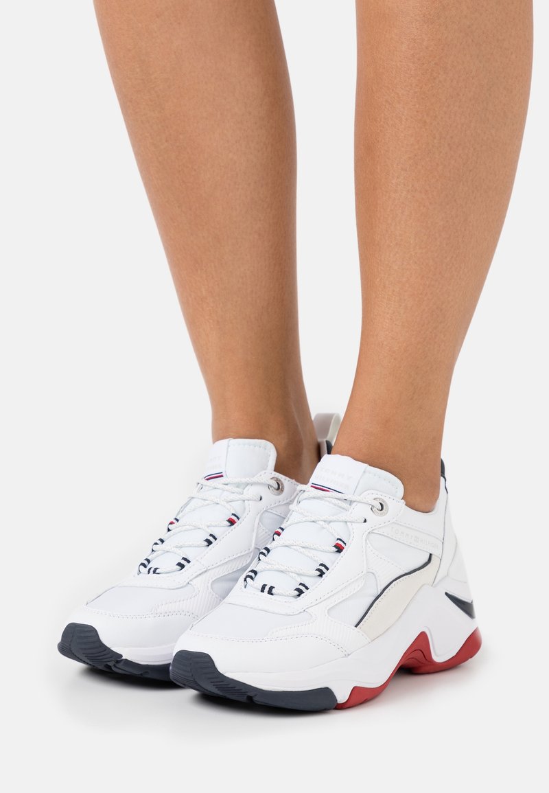 Baskets blanches avec une tige en cuir texturé, présentant des accents noirs et rouges, une semelle épaisse et un design classique à lacets.