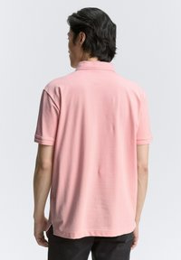 Rosa polo-shirt med krage, korta ärmar och rak fåll. Tyget verkar mjukt med en lätt lyster och minimala sömnadsdetaljer.