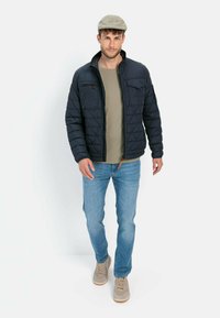 Granatowa pikowana kurtka z kołnierzem, dwoma przednimi kieszeniami i zapięciem na zamek, noszona na jasnoszarej koszuli, w połączeniu z niebieskimi dżinsami i beżowymi trampkami.