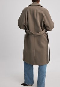 Pikk oliivroheline mantel vöödiga taljes, terava krae ja laia varrukaga, koos helesiniste laia säärega teksade ja mustade terava ninaga kingadega.