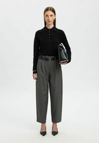 Camicia nera a maniche lunghe con bottoni, pantaloni grigi a vita alta e tacchi neri a punta. La modella tiene una borsa nera texturizzata.