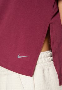 Camiseta deportiva burdeos de manga corta, con aberturas laterales y un logo de Nike en gris claro. La tela es suave y ligeramente texturizada.