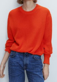Pull en tricot surdimensionné de couleur orange vif, avec un col rond et des poignets côtelés, associé à un jean en denim bleu.