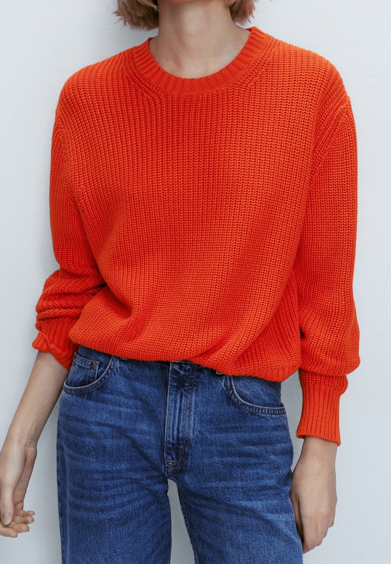Pull en tricot surdimensionné de couleur orange vif, avec un col rond et des poignets côtelés, associé à un jean en denim bleu.