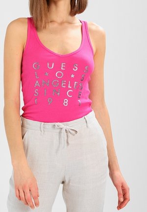 Femme portant un haut sans manches rose vif avec un texte argenté "Guess Los Angeles Since 1981" et un pantalon beige clair avec une ceinture nouée.
