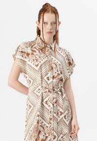 Abito a maniche corte in beige con un motivo astratto marrone. Presenta un colletto e una vita regolabile con lacci per una vestibilità personalizzabile, realizzato in tessuto leggero.