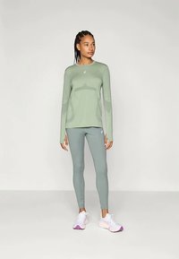 Gaisši zaļa garām piedurknēm sporta krekls ar rakstu, apvienots ar atbilstošām pieguļošām legingām. Apavi ir baltā krāsā ar rozā un violetiem akcentiem.