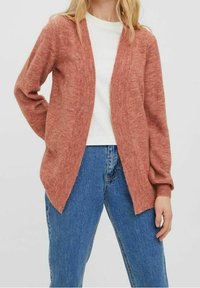 Rezavě zbarvený pletený kardigan s otevřeným předním dílem, dlouhými rukávy, žebrovanými manžetami a texturovaným povrchem, spárovaný s bílou košilí a modrými džínami.