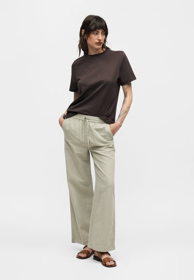 Γυναίκα με σκούρα μαλλιά που φοράει μαύρο κοντομάνικο, ανοιχτό μπεζ παντελόνι με φαρδιά πόδια και καφέ σανδάλια, στέκεται με τα χέρια στις τσέπες σε λευκό φόντο.