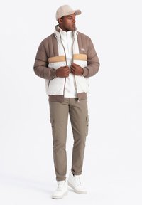Chaqueta de plumas marrón y blanca con una franja beige, combinada con una sudadera blanca con cremallera, pantalones cargo oliva y zapatillas deportivas blancas de caña alta.