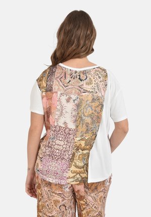 Donna con capelli castani lunghi che indossa una camicia bianca a maniche corte con patchwork paisley multicolore sulla schiena e pantaloni abbinati con motivo paisley.