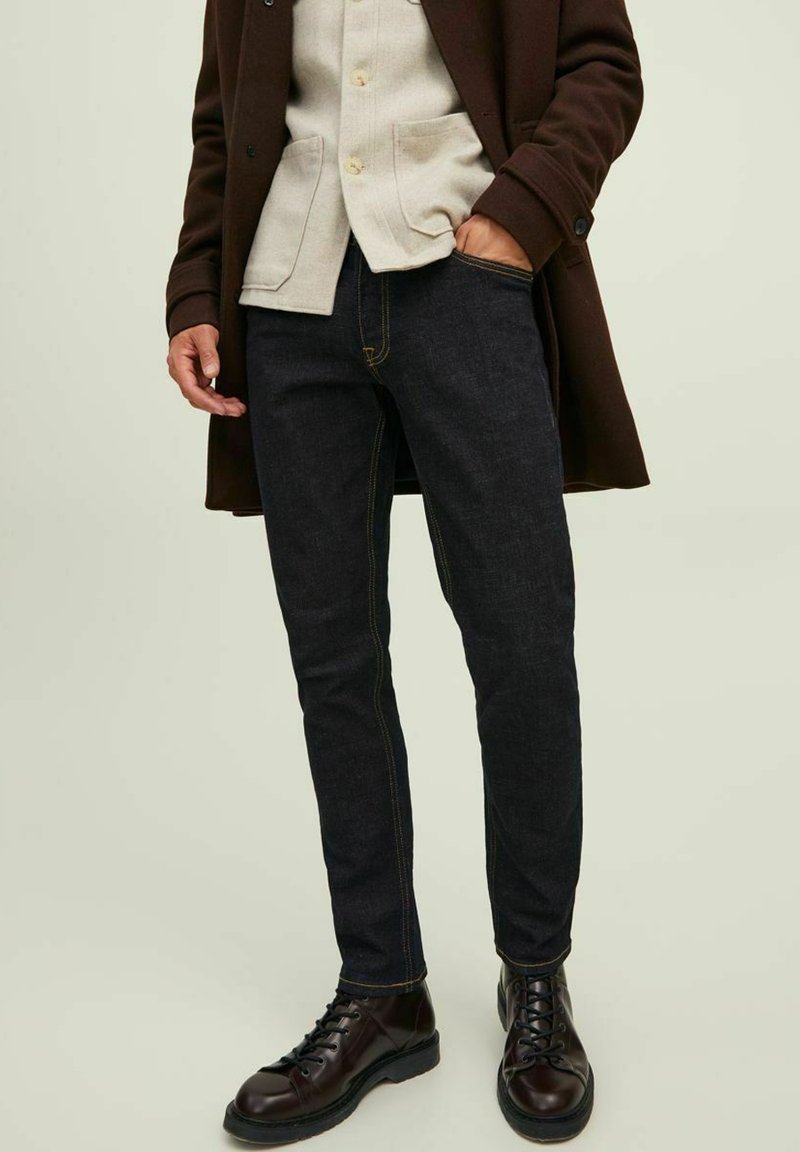 Jeans de ganga escuros com costura amarela contrastante, design slim-fit. Usados com uma camisa cinza clara e um casaco exterior castanho. Botas castanhas.