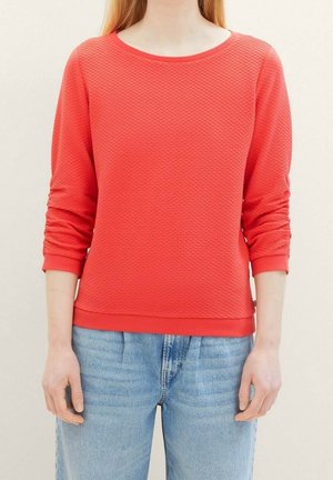 Frau trägt ein texturiertes korallenfarbenes Langarmoberteil mit gerafften Ärmeln und hellblaue, hoch taillierte Jeans, steht vor neutralem Hintergrund.