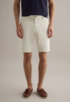 Hombre con pantalones cortos off-white hasta la rodilla, camisa azul marino y zapatos náuticos de cuero marrón, de pie contra un fondo neutro y sencillo.