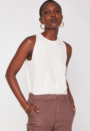 Ärmelloses weißes Stricktop mit hohem Kragen, kombiniert mit braunen elastischen Hosen mit taillenhohem Bund. Model trägt goldene Ohrstecker in Form von Ringen. Glatte Textur.