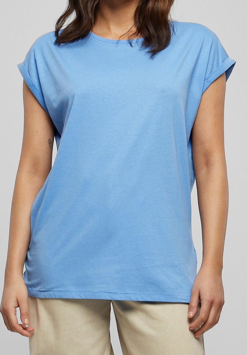 Femme portant un t-shirt ample bleu clair aux manches retroussées et un pantalon beige, debout devant un fond gris uni.