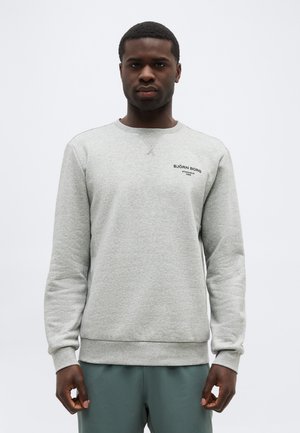 Mann trägt ein hellgraues Björn Borg Sweatshirt und grüne Hose, steht mit Blick nach vorne vor einem schlichten weißen Hintergrund.