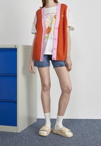 Henkilö, joka käyttää oranssia neulottua liiviä, jossa on vaaleanpunaista reunustusta, valkoista kukkakuosisia t-paitaa, farkkushortseja, valkoisia sukkia ja beigejä liukuvaatteisia sandaaleja, seisoo sinisen asiakirjahyllyn vieressä.