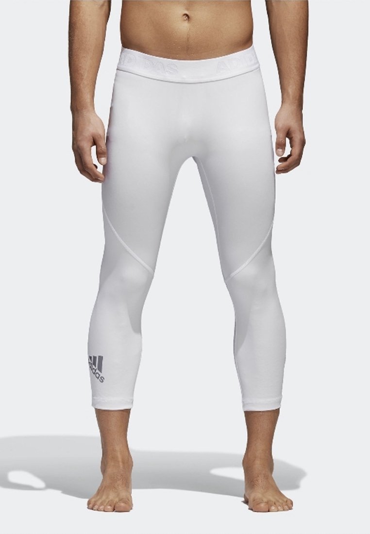 Homme portant des leggings de compression Adidas blancs coupés au-dessus des chevilles, debout pieds nus devant un fond uni.