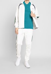 Vit zip-up jacka med gröna detaljer, turkos polo-shirt och vita sweatpants. Slät textur, atletisk design, med vita sneakers.