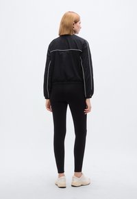 Μαύρο μπουφάν μπομμέρ με λευκές λεπτομέρειες, ελαστικά μανσέτες και ζωνάρι στη μέση, συνδυασμένο με μαύρα κολάν και ανοιχτόχρωμα αθλητικά παπούτσια.