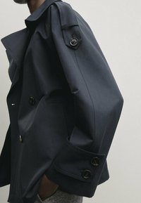 Granatowy płaszcz trench z podwójnym zapięciem, dużymi czarnymi guzikami oraz zbudowaną sylwetką z wyspecjalizowanymi akcentami na ramionach.