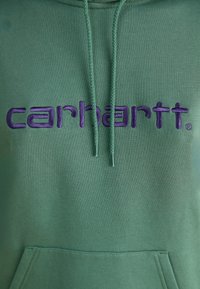 Hanorac verde cu buzunar frontal tip kangaroo, glugă cu șnur și logo-ul brodat „carhartt” în culoarea purpurie pe piept. Textură moale.