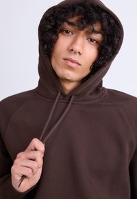 Brown hoodie with a drawstring hood, featuring raglan sleeves and soft texture. Model holds the drawstring, displaying a relaxed fit.