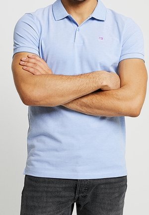 Homme portant un polo bleu clair et un jean noir, debout les bras croisés devant un fond uni.