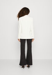 Bílý strukturovaný blazer s dlouhými rukávy a knoflíkovými manžetami, kombinovaný s černými rozšířenými kalhotami, na světlém jednobarevném pozadí.